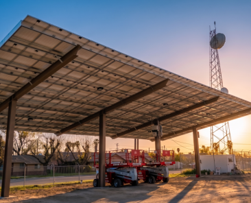 A large solar panel canopy, part of ongoing municipal solar projects, is installed over a dirt lot at sunset. Two scissor lifts are parked underneath, with trees, houses, a utility tower, and a fence in the background.