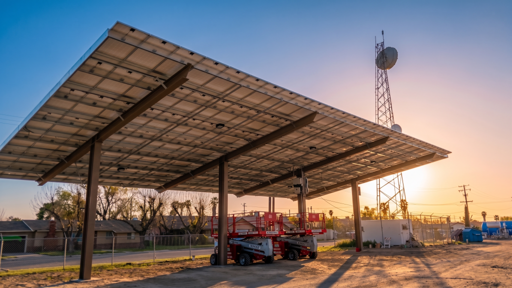 A large solar panel canopy, part of ongoing municipal solar projects, is installed over a dirt lot at sunset. Two scissor lifts are parked underneath, with trees, houses, a utility tower, and a fence in the background. A large solar panel canopy, part of ongoing municipal solar projects, is installed over a dirt lot at sunset. Two scissor lifts are parked underneath, with trees, houses, a utility tower, and a fence in the background.
