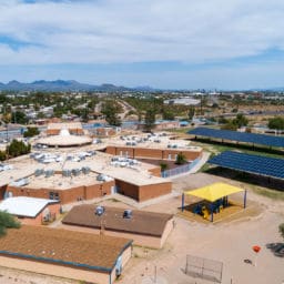 Aerial view of a school campus with solar panels on covered structures, highlighting solar PPA for schools, surrounded by desert landscape, scattered buildings, and sports areas. Mountains and cityscape are visible in the background under a partly cloudy sky.