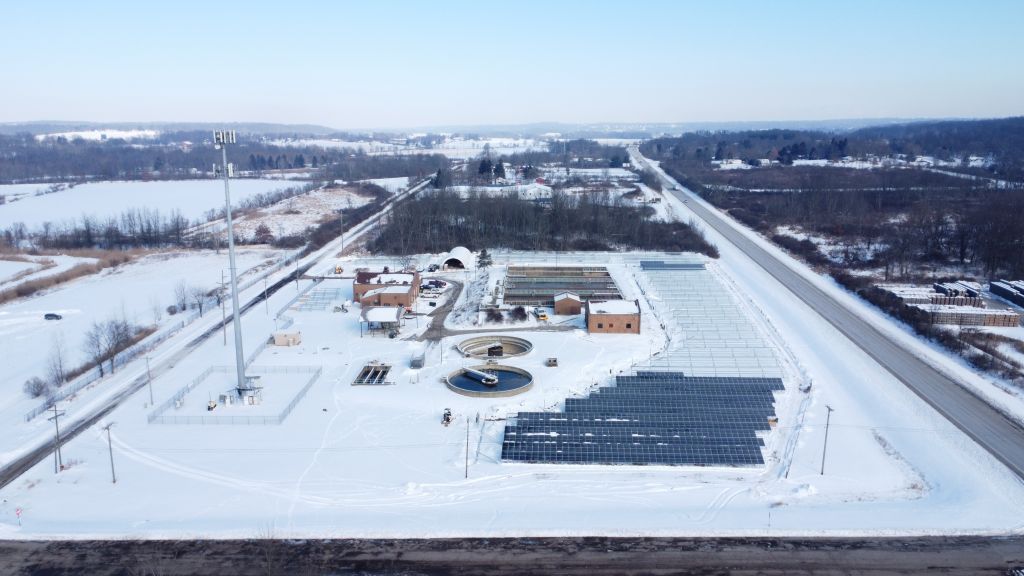 Aerial view of a snow-covered water treatment facility with commercial solar financing, solar panels, buildings, and a tall communications tower, surrounded by fields and roads in a rural winter landscape.