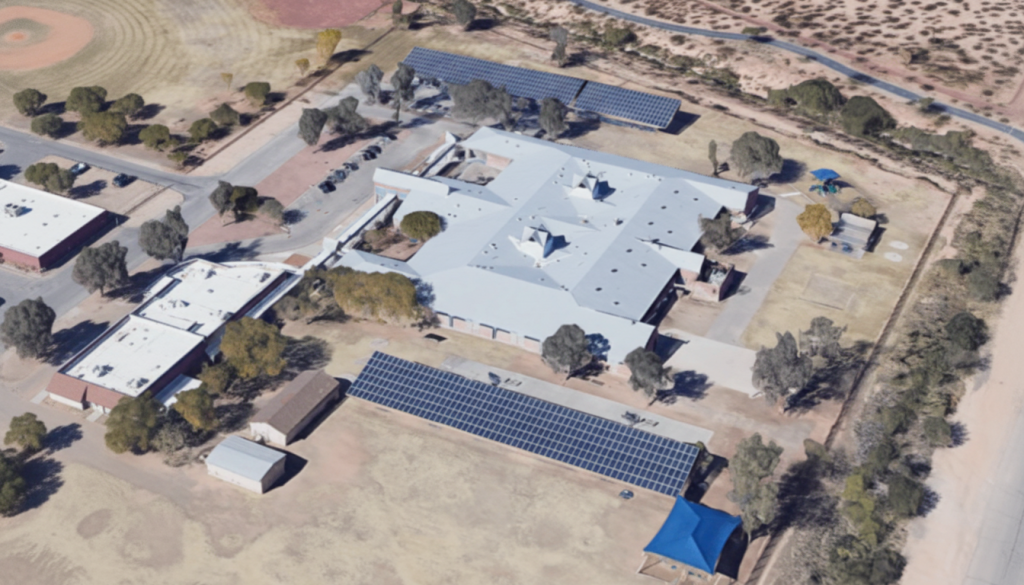Aerial view of a large building complex with white roofs, surrounded by trees and dirt lots, featuring two long rows of solar panels on the ground—ideal for commercial solar financing or adding EV charging for businesses.