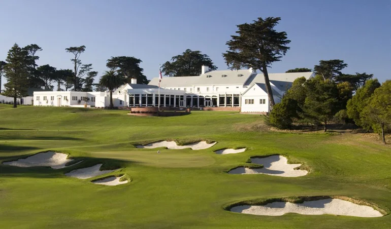 A large white clubhouse at the Cal Club of South San Francisco sits behind a well-maintained golf course with sand bunkers, green grass, and surrounding trees under a clear blue sky—a setting enhanced by its 486 kW energy system.