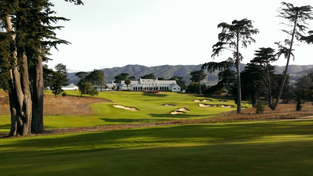 A scenic golf course with manicured fairways, several sand bunkers, and tall trees, set against hills and a large white clubhouse that could benefit from commercial solar PPA solutions.
