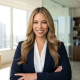 A woman with long, wavy blonde hair, wearing a navy blazer and white blouse, stands smiling with arms crossed in a bright, modern office inspired by Steven Campbell, featuring large windows and bookshelves in the background.
