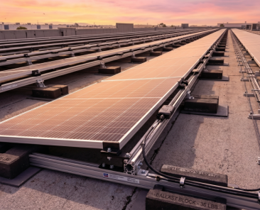 Rows of solar panels, part of a commercial solar project, are installed on a flat rooftop at sunset. Conduits and ballast blocks secure the system as the panels reflect the warm, pinkish glow of the sky.